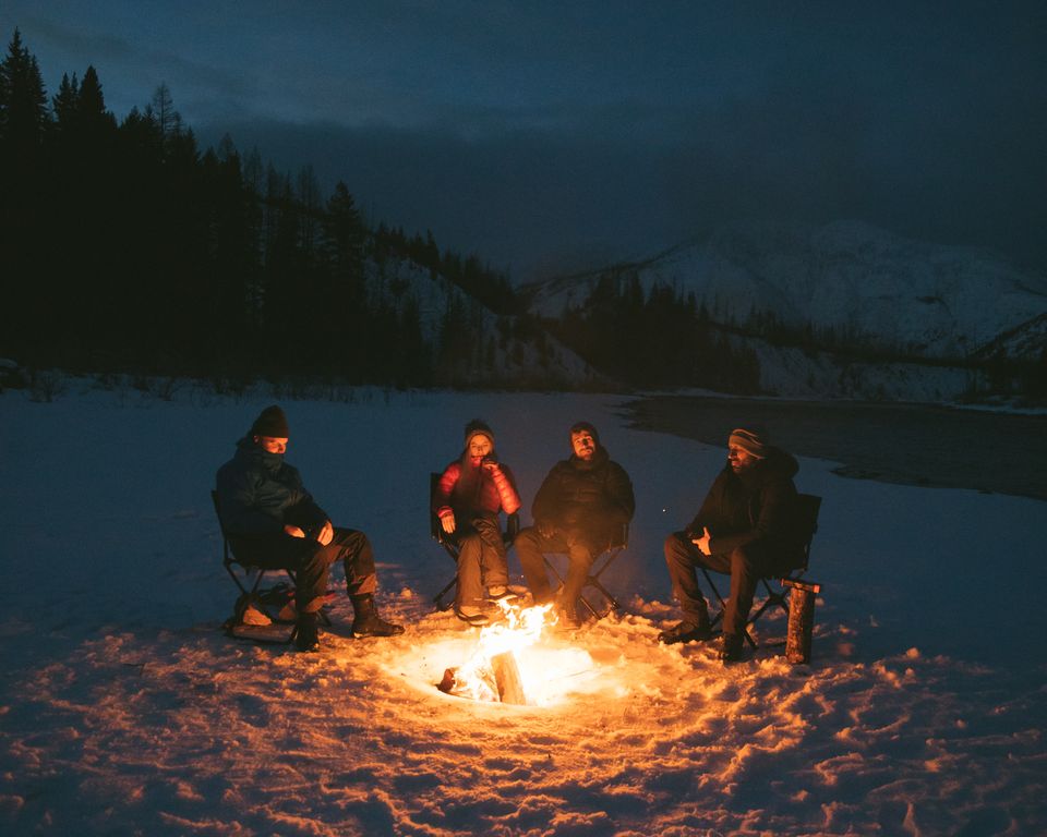 Four friends sit around a campfire on the snow at dusk