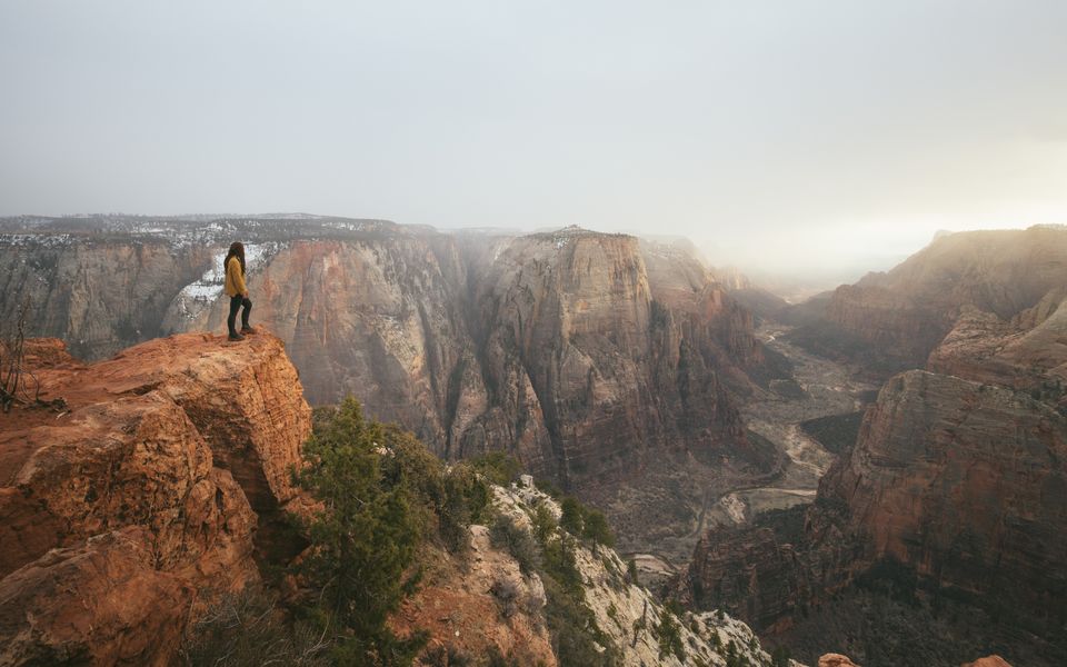 A person stands on a canyon rim above a vast red-rock valley