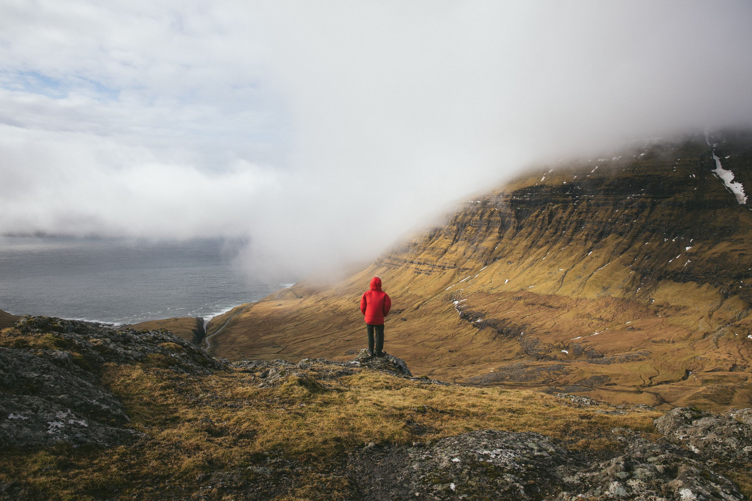A hiker in a red jacket stands on a grassy ridge under rolling cloud