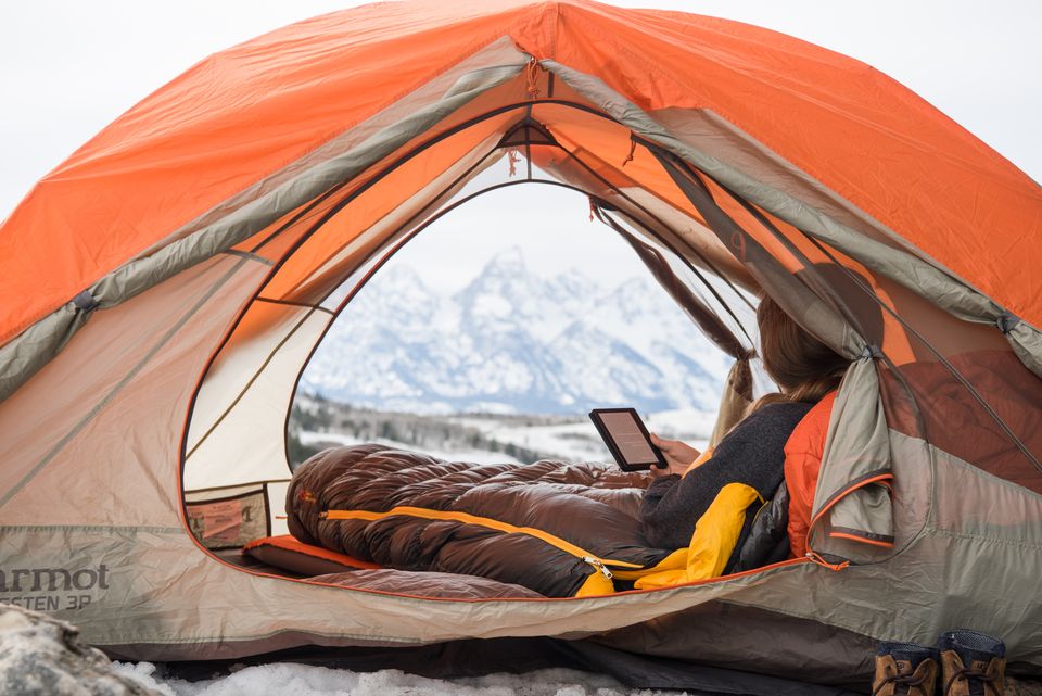 A woman reads in an orange tent with snowy mountains outside