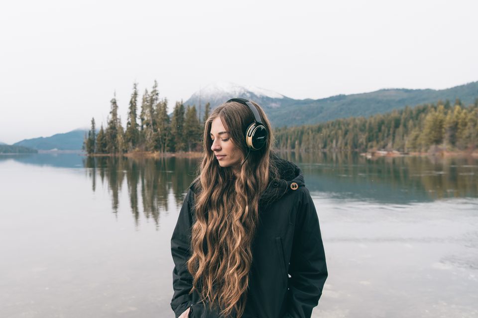 A woman in headphones and a dark parka by a lake with a small island
