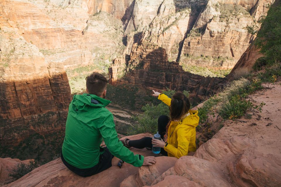 Two hikers in bright jackets resting on a red rock ledge above a deep canyon