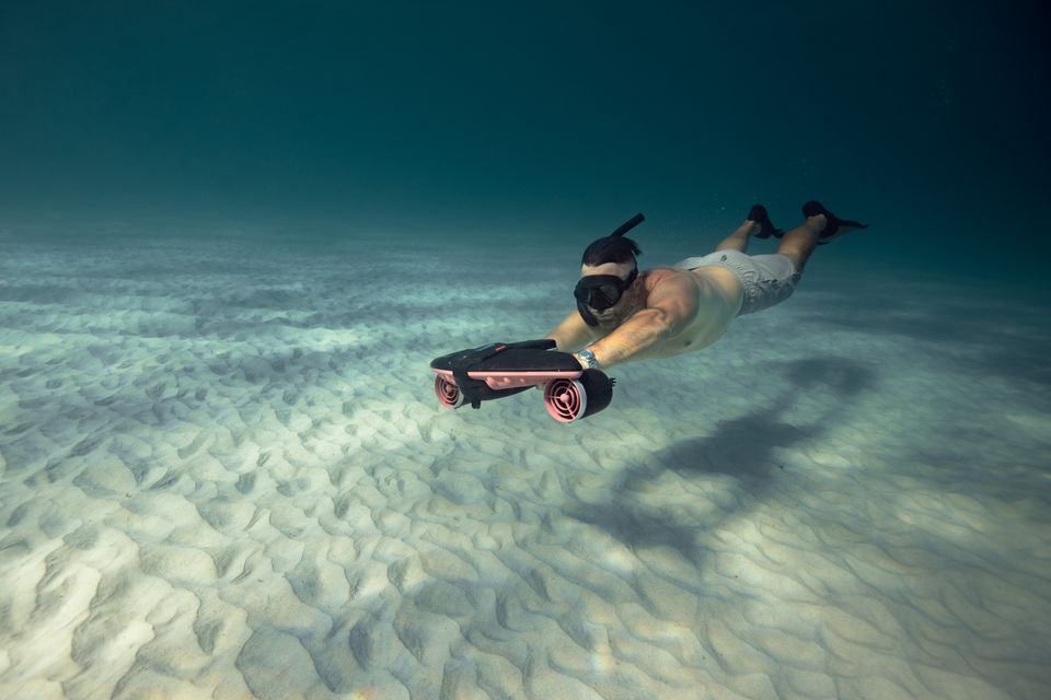 A diver glides over a sandy seafloor holding an underwater scooter