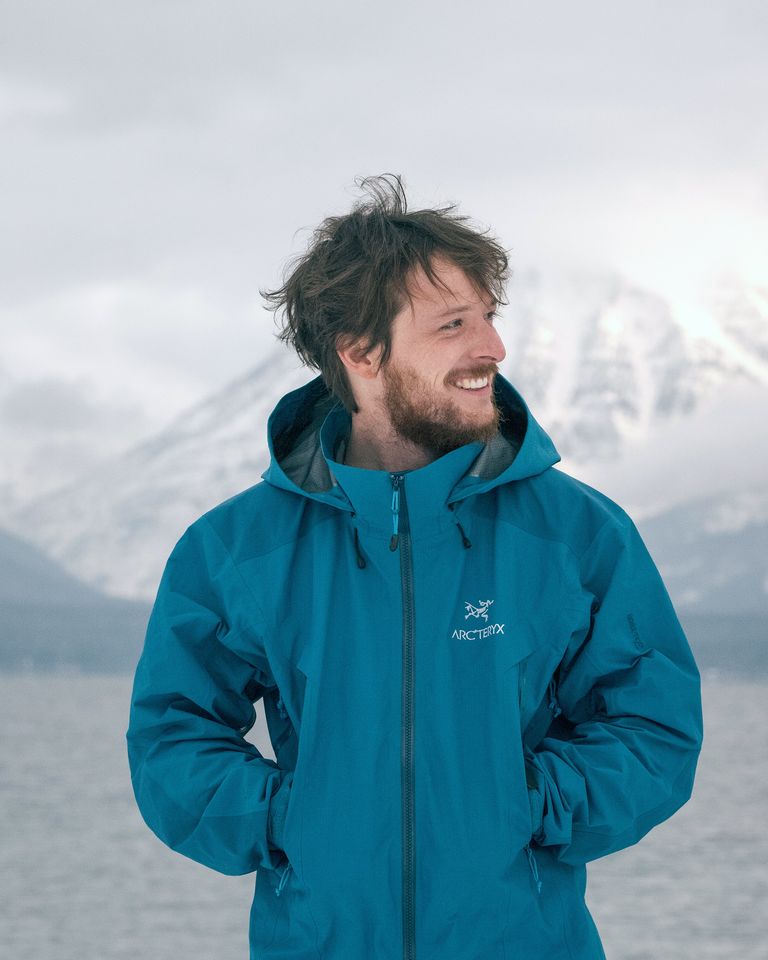 Morgan Phillips in a teal rain jacket, smiling, with a lake and snowy mountains behind him