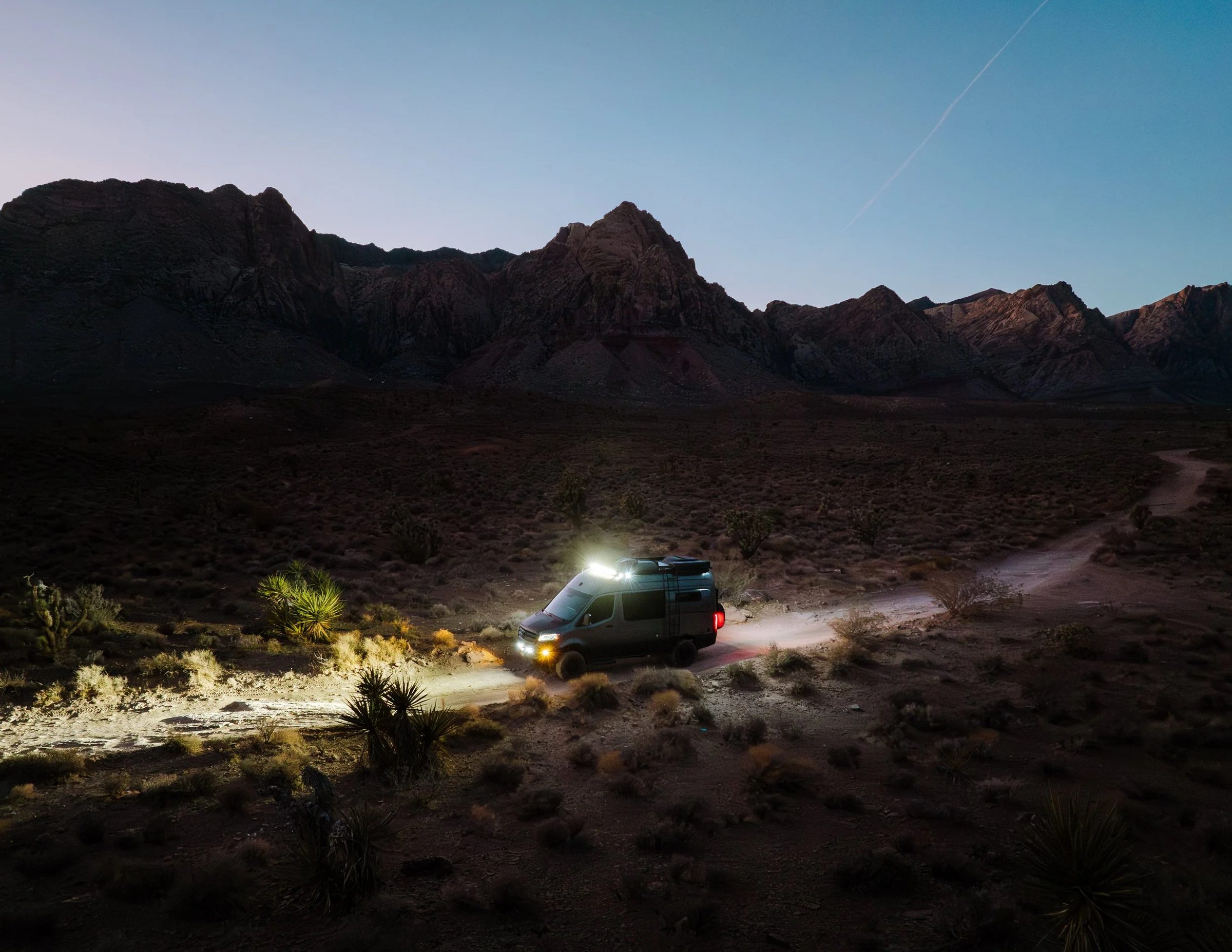 A camper van with its lights on, parked on a desert dirt road below dark mountains at dusk